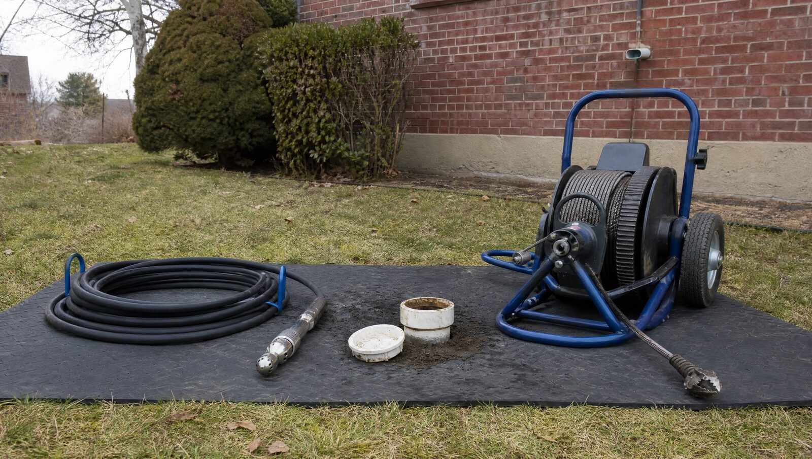 A jetter hose with a rotating nozzle and a drum cable machine with coiled steel snake cable laid side by side on a mat beside an open sewer cleanout.