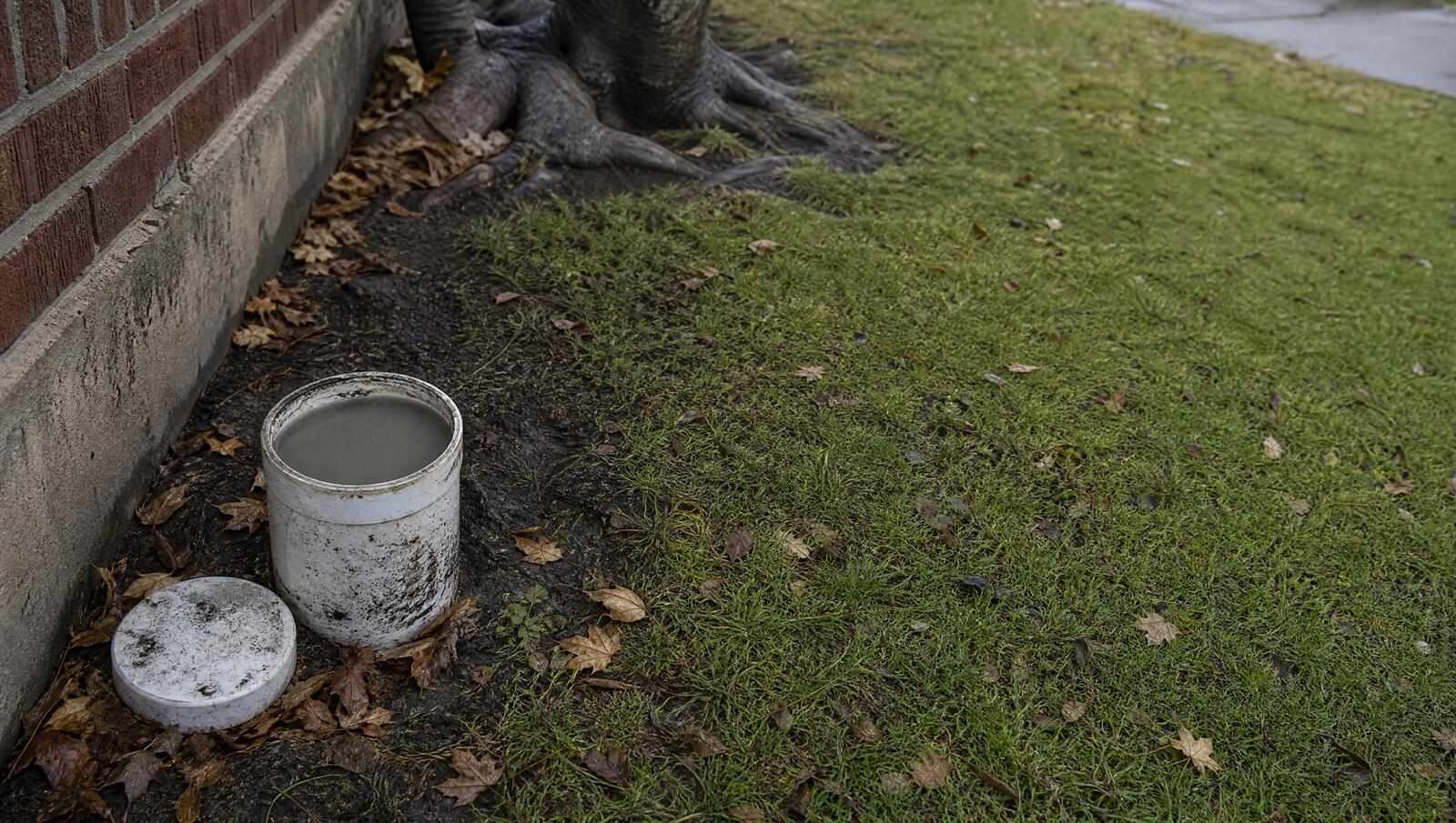 Opened sewer cleanout riser beside the brick foundation of a Denver bungalow, cap set aside, standing cloudy liquid sitting high in the pipe where a blockage has stopped it draining.