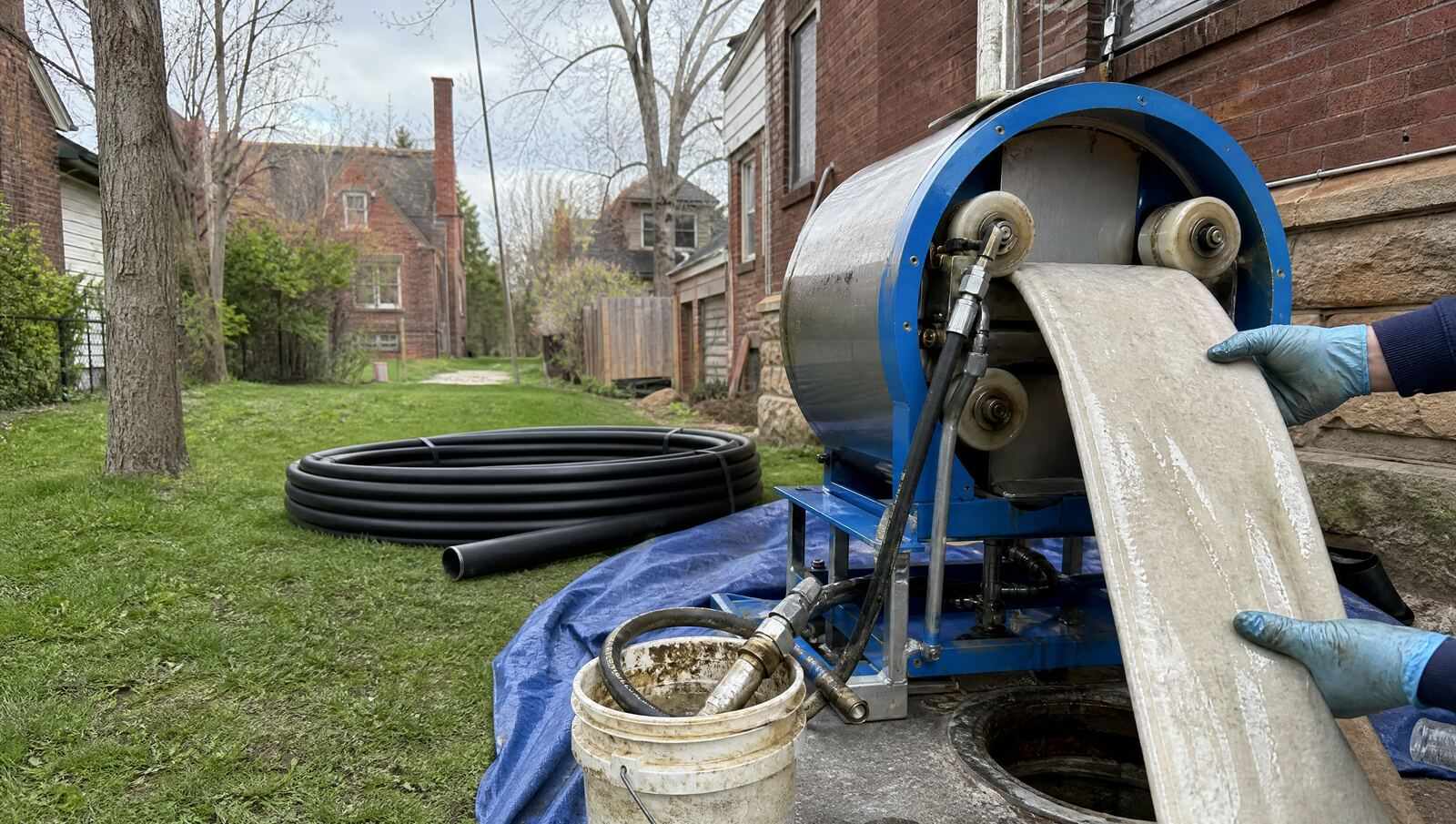 A resin saturated liner being fed into an inversion drum over a sewer access in a Denver side yard, with a coil of new HDPE replacement pipe lying on the grass alongside.