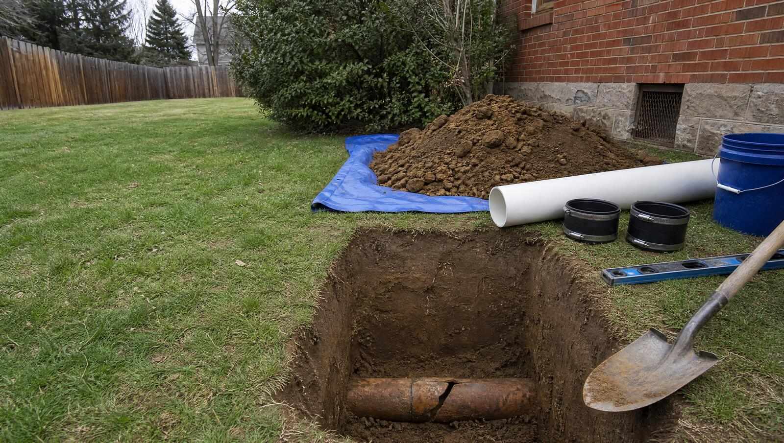 A cracked section of clay sewer pipe exposed at the bottom of a compact spot repair trench in a Denver side yard, with replacement pipe and repair couplings staged at the trench edge.